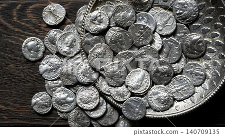 Top view of ancient Roman and Greek silver coins spilling from an ornate metal dish onto a dark wooden table. Historical money collection and antique treasure concept 140709135