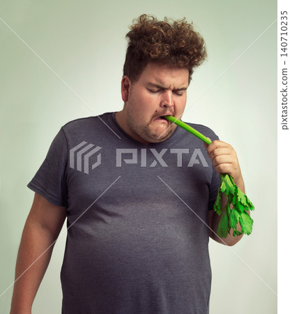 Diet, plus size and unhappy man eating vegetable for health benefit, nutrition and wellness in studio by white background. Weight loss, celery and male person for healthy food, wellbeing and detox Diet, plus size and unhappy man eating vegetable for health benefit, nutrition and wellness in studio by white background. Weight loss, celery and male person for healthy food, wellbeing and detox 140710235
