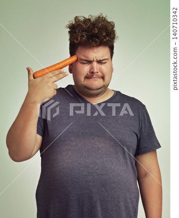 Depression, vegetable and hand gun with plus size man in studio on gray background for diet, health or nutrition. Sad, unhappy and carrot to head of young person in conflict with vegetarian food Depression, vegetable and hand gun with plus size man in studio on gray background for diet, health or nutrition. Sad, unhappy and carrot to head of young person in conflict with vegetarian food 140710342