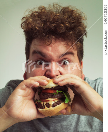 Takeaway, burger and food for man in studio with meal for big eater, plus size or greed. Young person with meat in mouth for hunger, silly and bite indoor for delicious, taste and sauce for portrait 140710392