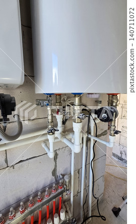 Domestic heating system with gas boiler, piping, and manifold on concrete wall in utility room. Domestic heating system with gas boiler, piping, and manifold on concrete wall in utility room. 140711072