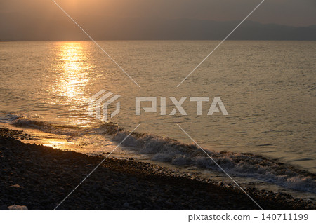 Sunset casts golden reflections over serene waters and pebbled shoreline in a tranquil coastal setting 140711199