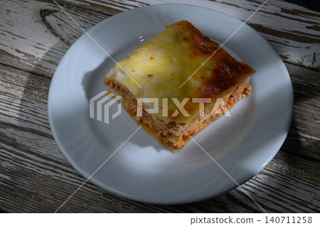 Delicious homemade lasagna served on a white plate against a rustic wooden background Delicious homemade lasagna served on a white plate against a rustic wooden background 140711258