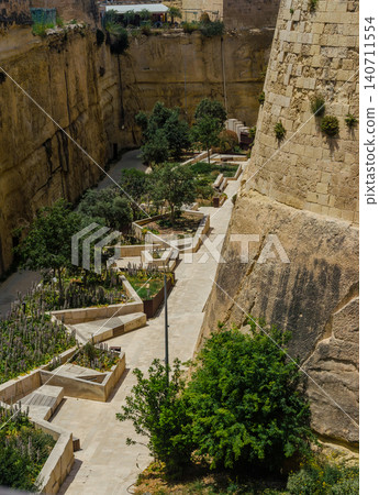 Landscaped dry moat ditch garden below massive Valletta fortification walls Malta with terraced planters tree pathways and bastion Landscaped dry moat ditch garden below massive Valletta fortification walls Malta with terraced planters tree pathways and bastion 140711554