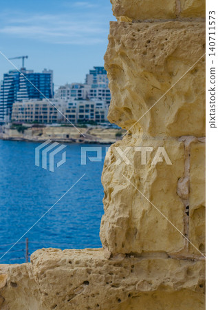 Golden limestone wall texture in focus with blurred Sliema Malta coastal city skyline and blue Mediterranean sea in background 140711573