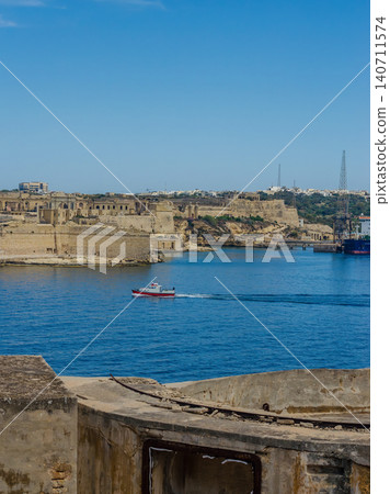 Grand Harbour Valletta Malta with red boat on blue sea historic fortifications limestone forts and old gun emplacement foreground 140711574