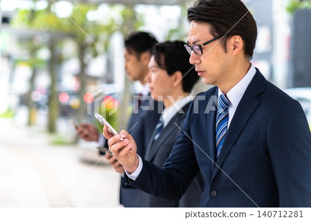 A man in a suit is using his smartphone while waiting for a bus at a bus stop. A man in a suit is using his smartphone while waiting for a bus at a bus stop. 140712281