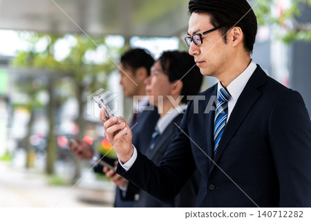 A man in a suit is using his smartphone while waiting for a bus at a bus stop. A man in a suit is using his smartphone while waiting for a bus at a bus stop. 140712282