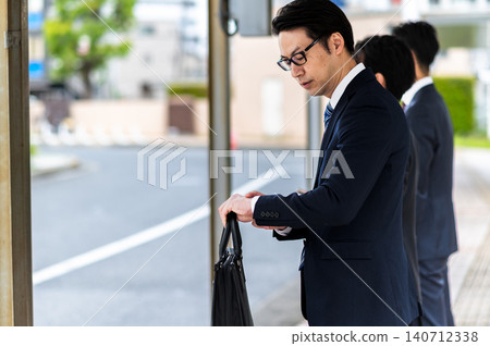 A man in a suit checks his watch while waiting for a bus at a bus stop. 140712338