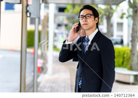 A man in a suit is talking on the phone while waiting for a bus at a bus stop. A man in a suit is talking on the phone while waiting for a bus at a bus stop. 140712364