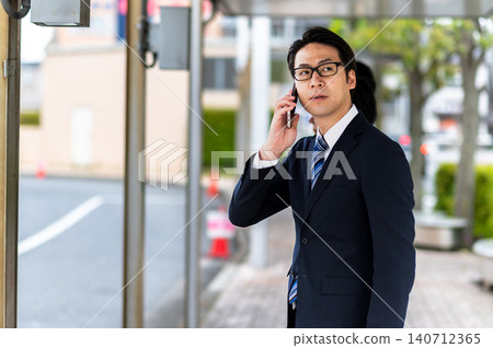 一名身穿西裝的男子在公車站等車時正在打電話。 140712365