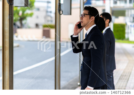 A man in a suit is talking on the phone while waiting for a bus at a bus stop. A man in a suit is talking on the phone while waiting for a bus at a bus stop. 140712366