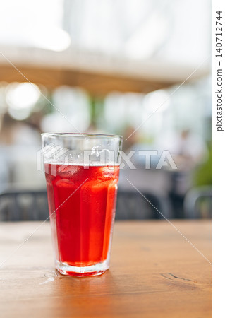 Tall clear glass filled with refreshing red iced beverage or cold brew tea standing on wooden table in outdoor summer cafe with blurry bokeh background 140712744