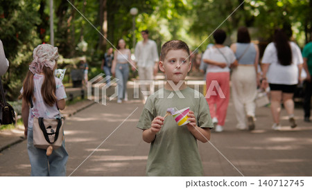 Boy in olive green t-shirt holds colorful paper airplane while standing on shaded pedestrian path in leafy urban park; blurred walkers and couples move behind Boy in olive green t-shirt holds colorful paper airplane while standing on shaded pedestrian path in leafy urban park; blurred walkers and couples move behind 140712745