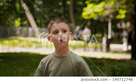 Close-up portrait of young boy standing in sunlit city park, wearing olive green shirt, with short brown hair, wide eyes, open mouth, and surprised face Close-up portrait of young boy standing in sunlit city park, wearing olive green shirt, with short brown hair, wide eyes, open mouth, and surprised face 140712746