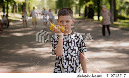 Young boy holding corn on cob to mouth while standing on paved park walkway surrounded by tall trees and dappled sunlight, casual patterned t-shirt, relaxed Young boy holding corn on cob to mouth while standing on paved park walkway surrounded by tall trees and dappled sunlight, casual patterned t-shirt, relaxed 140712747