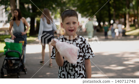 Young boy stands on tree-lined park pathway holding pink cotton candy while smiling toward camera; casual summer clothing, patterned T-shirt, short haircut Young boy stands on tree-lined park pathway holding pink cotton candy while smiling toward camera; casual summer clothing, patterned T-shirt, short haircut 140712748