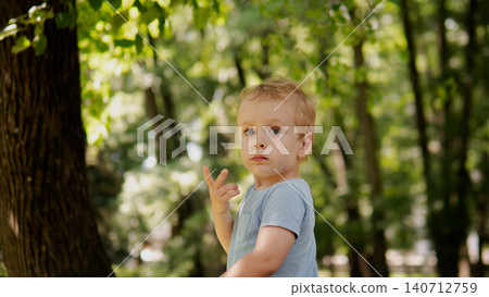 Toddler standing among tall trees in leafy forest during daytime, wearing light blue short sleeve shirt and light pants, fair hair and curious facial Toddler standing among tall trees in leafy forest during daytime, wearing light blue short sleeve shirt and light pants, fair hair and curious facial 140712759