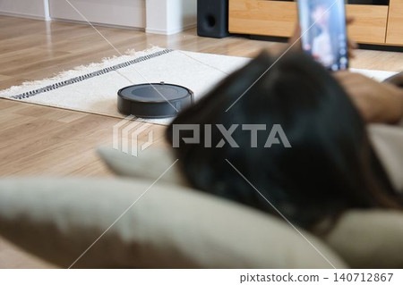 一位家庭主婦正躺在沙發上放鬆,旁邊放著一台掃地機器人。 一位家庭主婦正躺在沙發上放鬆,旁邊放著一台掃地機器人。 140712867