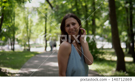 Young woman wearing light blue sleeveless top stands on shaded tree-lined park path, smiling at camera while touching hair; dappled sunlight filters through Young woman wearing light blue sleeveless top stands on shaded tree-lined park path, smiling at camera while touching hair; dappled sunlight filters through 140712869