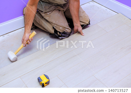 Contractor kneels on laminate floor lays down new planks while using hammer, measuring tape in room renovation Contractor kneels on laminate floor lays down new planks while using hammer, measuring tape in room renovation 140713527