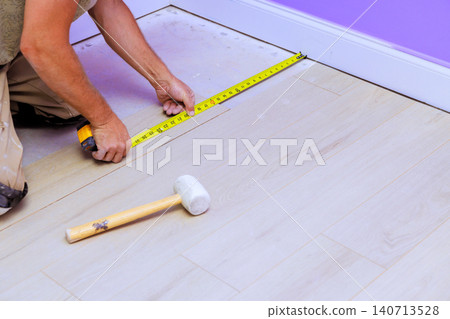 Handy worker is measuring wooden floor while preparing to lay new flooring in room with purple walls Handy worker is measuring wooden floor while preparing to lay new flooring in room with purple walls 140713528