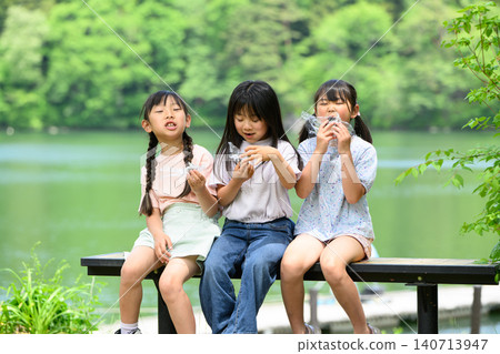一群小學生在湖邊長椅上,被綠樹環繞,開心地一起吃著飯糰。 一群小學生在湖邊長椅上,被綠樹環繞,開心地一起吃著飯糰。 140713947