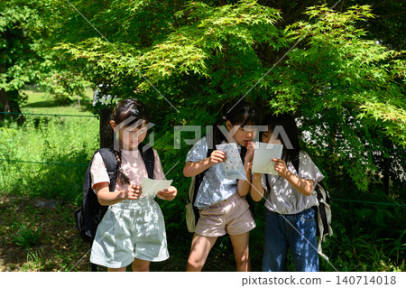 Elementary school girls look at a map, find a route, and point it out amidst the lush greenery of nature. Elementary school girls look at a map, find a route, and point it out amidst the lush greenery of nature. 140714018