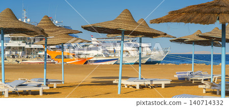 Hurghada, Egypt - Mar 3 2024, panoramic view of the beach with sun loungers and straw umbrellas, without people, moored yachts and boats are visible in the background, at daytime, Hurghada, Egypt Hurghada, Egypt - Mar 3 2024, panoramic view of the beach with sun loungers and straw umbrellas, without people, moored yachts and boats are visible in the background, at daytime, Hurghada, Egypt 140714332
