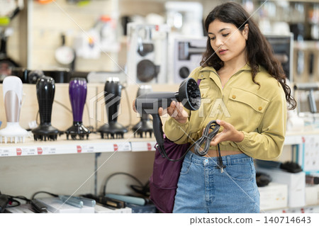 Latina woman stands in a store with household appliances and carefully chooses a hairdryer Latina woman stands in a store with household appliances and carefully chooses a hairdryer 140714643