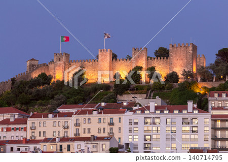 Illuminated medieval fortress walls rise above the rooftops of Lisbon during blue hour. Illuminated medieval fortress walls rise above the rooftops of Lisbon during blue hour. 140714795