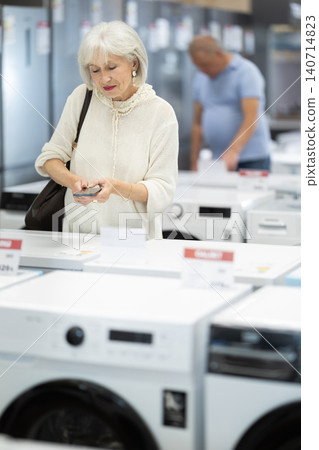 Elderly woman choosing washing machine scans QR code on the price tag to check technical specifications Elderly woman choosing washing machine scans QR code on the price tag to check technical specifications 140714823