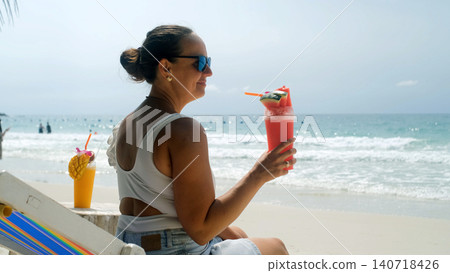 Young woman wearing sunglasses sipping a refreshing watermelon smoothie while sitting on a beach chair with sea and sky in background. Tropical relaxation concept Young woman wearing sunglasses sipping a refreshing watermelon smoothie while sitting on a beach chair with sea and sky in background. Tropical relaxation concept 140718426