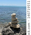Balanced stones on rocky seashore photo with calm blue sea, clear sky and distant sailboat, peaceful coastal landscape for travel, summer vacation, meditation, balance, nature and wellness design 140719551
