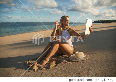 Woman sits on sand and waves while video chatting 140719679