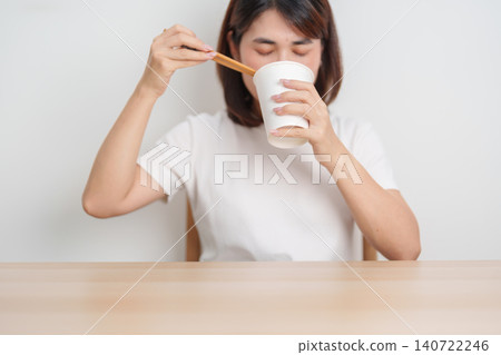 Happy young Asian woman enjoying a delicious cup of hot instant noodles while relaxing at home, Cheerful female eating instant ramen with a smile at apartment. Simple lifestyle and quick meal concept 140722246