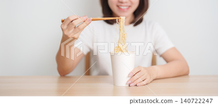Happy young Asian woman enjoying a delicious cup of hot instant noodles while relaxing at home, Cheerful female eating instant ramen with a smile at apartment. Simple lifestyle and quick meal concept Happy young Asian woman enjoying a delicious cup of hot instant noodles while relaxing at home, Cheerful female eating instant ramen with a smile at apartment. Simple lifestyle and quick meal concept 140722247
