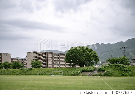 Under a cloudy sky, a view of apartment buildings and mountain ranges stands in the distance behind a lush green field and large trees. Under a cloudy sky, a view of apartment buildings and mountain ranges stands in the distance behind a lush green field and large trees. 140722639
