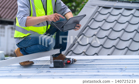 Roofer in Safety Vest Using Tablet on Metal Roof During Construction Inspection Roofer in Safety Vest Using Tablet on Metal Roof During Construction Inspection 140725219