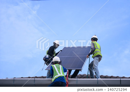 Three Workers Installing Solar Panel on Rooftop Against Blue Sky 140725294