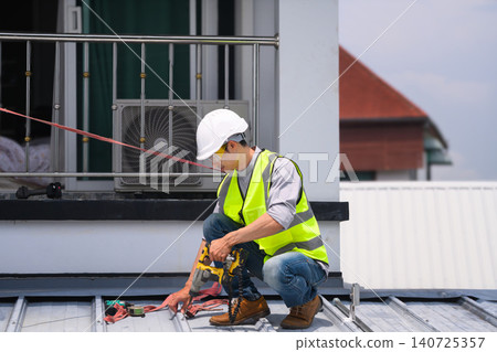 Asian Male Roofer Using Power Drill on Metal Roof Installation with Safety Equipment Outdoors Asian Male Roofer Using Power Drill on Metal Roof Installation with Safety Equipment Outdoors 140725357
