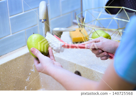 A young vegan washes garden apples under the tap in a sunny kitchen, brushing each one thoroughly while practicing safe food habits. A young vegan washes garden apples under the tap in a sunny kitchen, brushing each one thoroughly while practicing safe food habits. 140725568