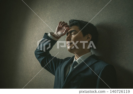 一個身穿西裝的男人靠在牆上,一手扶著額頭。 一個身穿西裝的男人靠在牆上,一手扶著額頭。 140726119