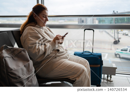 Young woman traveler sitting in airport waiting area and using smartphone Young woman traveler sitting in airport waiting area and using smartphone 140726534