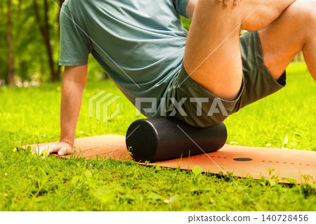 Man doing myofascial release with a foam roller on a mat in a green park. Postworkout recovery, massage, and therapy for muscle tension. Man doing myofascial release with a foam roller on a mat in a green park. Postworkout recovery, massage, and therapy for muscle tension. 140728456