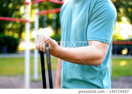 Close up of a man's arm doing a fitness workout with a resistance band. Outdoor sport training and exercise in a sunny summer park. Close up of a man's arm doing a fitness workout with a resistance band. Outdoor sport training and exercise in a sunny summer park. 140728487