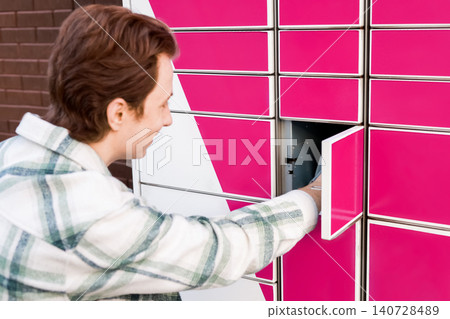 Smiling man picking up a package from an automated parcel locker. Convenient self-service terminal for e-commerce, online shopping, and contactless delivery logistics. Smiling man picking up a package from an automated parcel locker. Convenient self-service terminal for e-commerce, online shopping, and contactless delivery logistics. 140728489