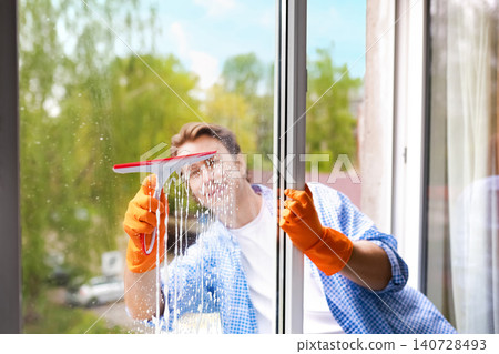 Smiling man doing housework by washing a glass window with a squeegee and soapy water. Male cleaner wearing orange gloves performing a cleaning chore. 140728493