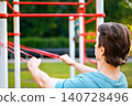 Active man doing a fitness workout in an outdoor park. He is performing an exercise for strength training using a red resistance band on a street gym structure. 140728496
