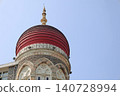 A low-angle shot featuring the iconic red dome of the Taj Mahal Palace Hotel in Mumbai showcasing its intricate Indo-Saracenic architectural details against a clear pale blue sky. 140728994